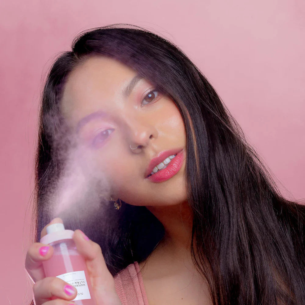 Woman holding a pink bottle with a spray mist over her face against a pink background